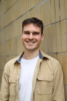 Junger Mann lehnt an gelber Mauer, Gesicht pixelisiert, beige Cordjacke und Weißes T‑Shirt.